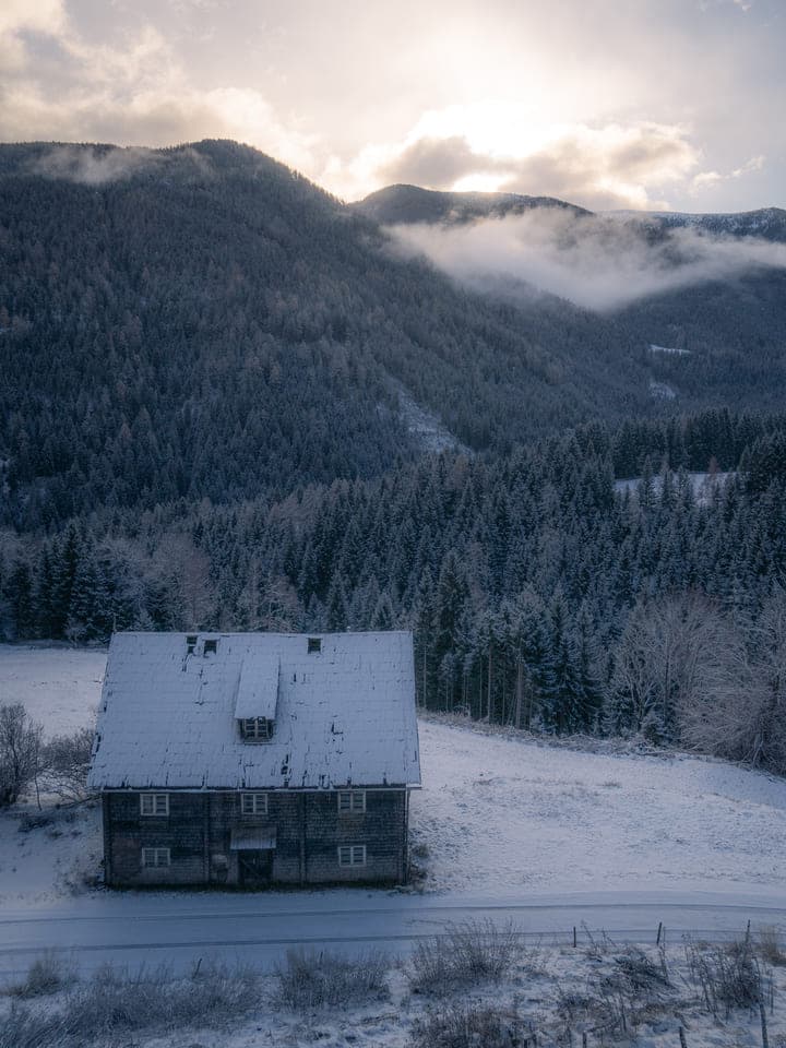 Snow-covered cabin below a forested mountain slope